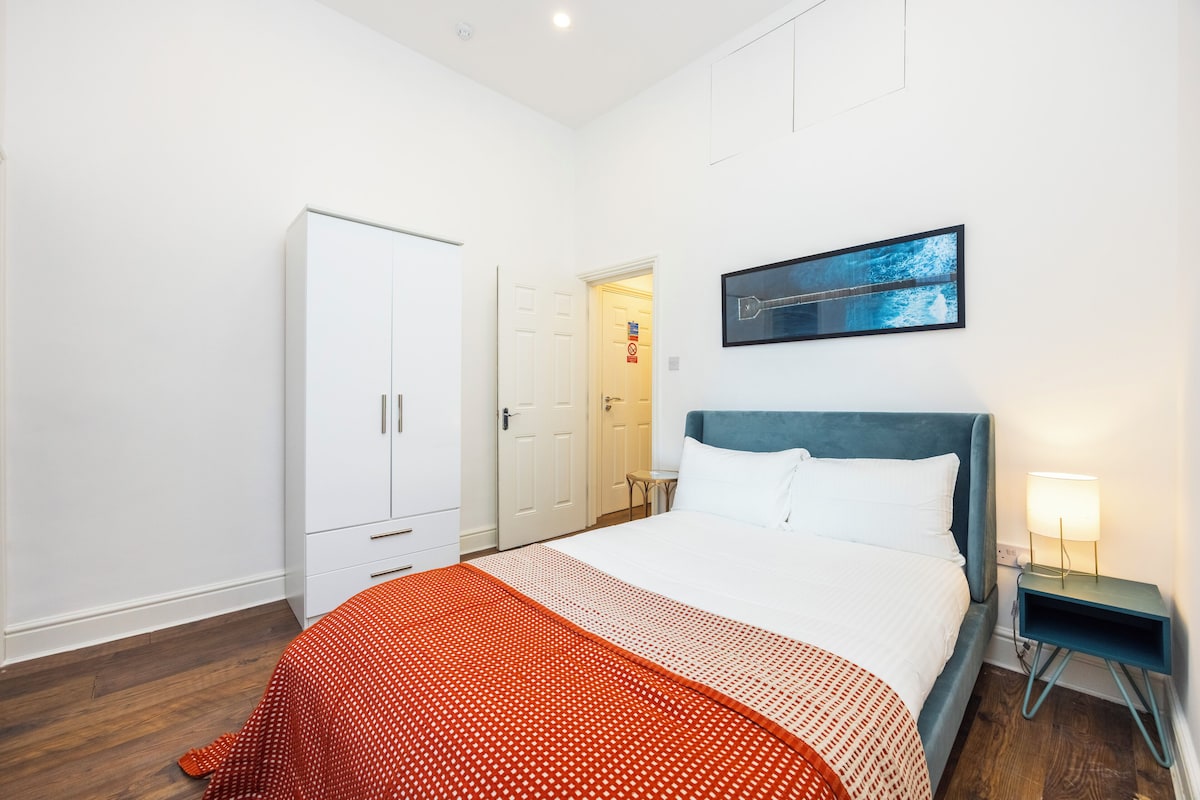 A hotel room with a bed, a wardrobe, a desk with a lamp, and a painting on the wall.