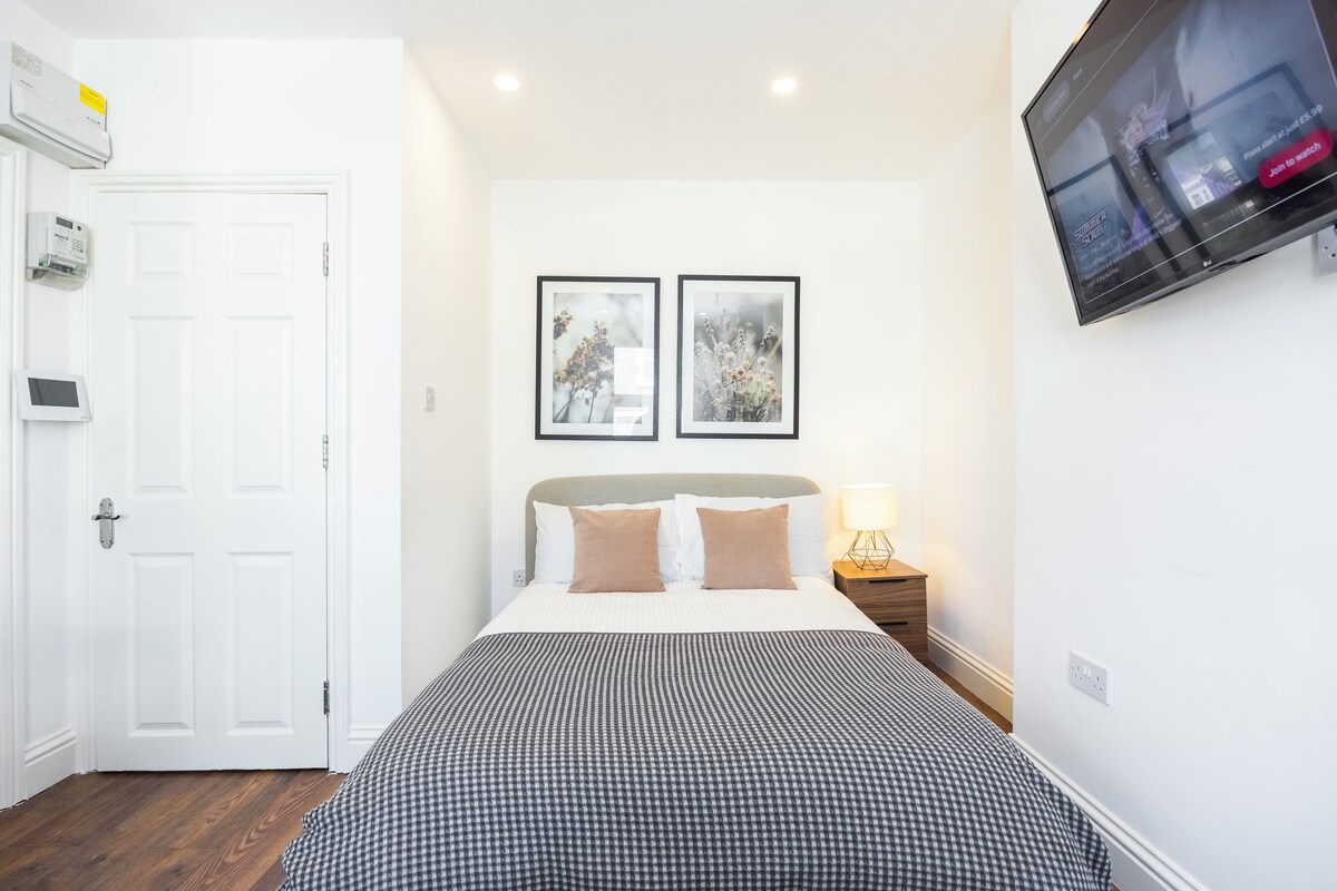 A hotel room with a bed, a bedside table with a lamp, a mounted TV, and a door with a lock.