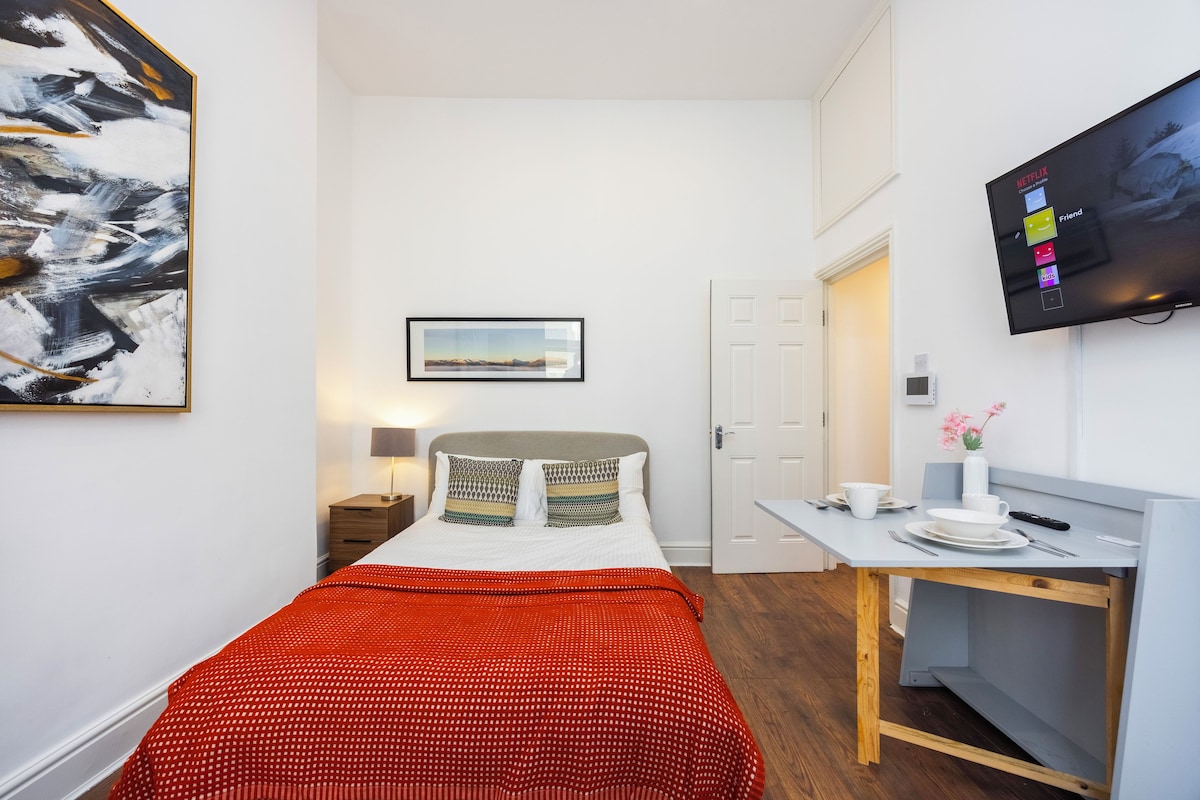 A hotel room with a bed, a desk, a television, and a painting on the wall.