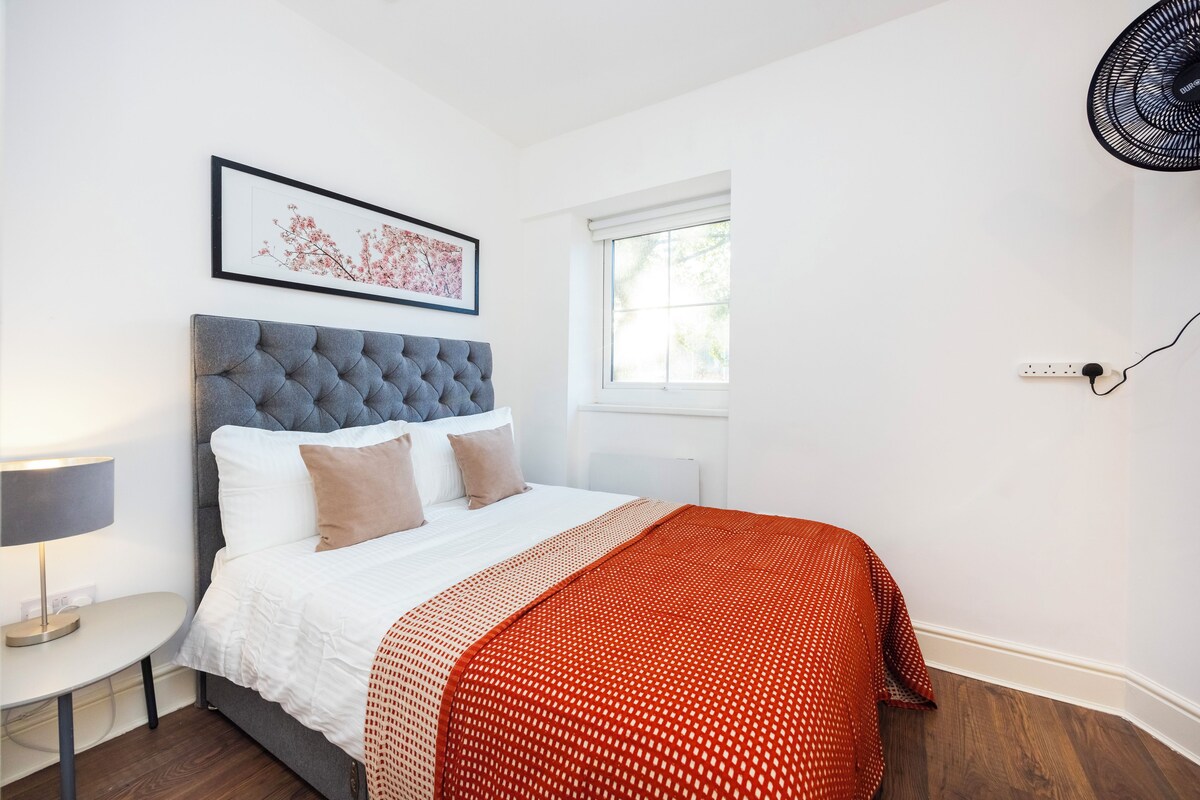 A bedroom with a bed, a bedside table, a lamp, a window, and a power outlet.