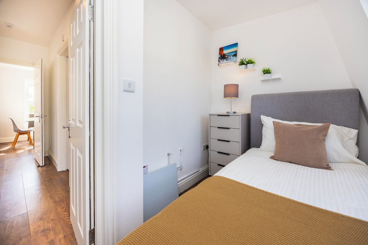A bedroom with a bed, bedside table, lamp, and a wall-mounted shelf with plants.