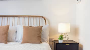 A neatly made bed with a striped comforter and two pillows, a bedside table with a lamp and a small plant, and a wall-mounted power outlet.