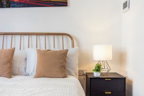 A neatly made bed with a striped comforter and two pillows, a bedside table with a lamp and a small plant, and a wall-mounted power outlet. - Earl's Court Residence (London)