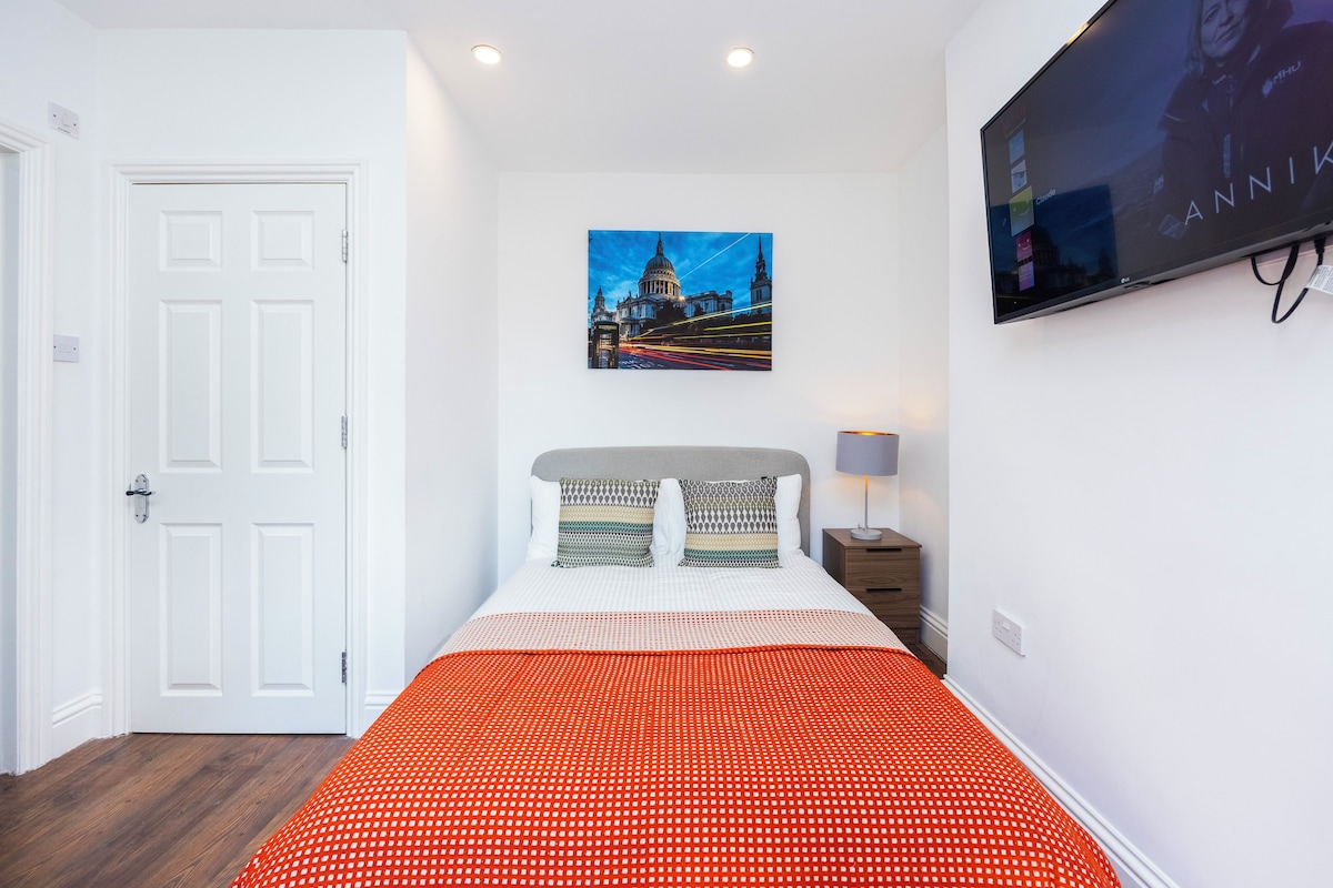 A bedroom with a bed, a nightstand, a lamp, a wall-mounted TV, and a door.