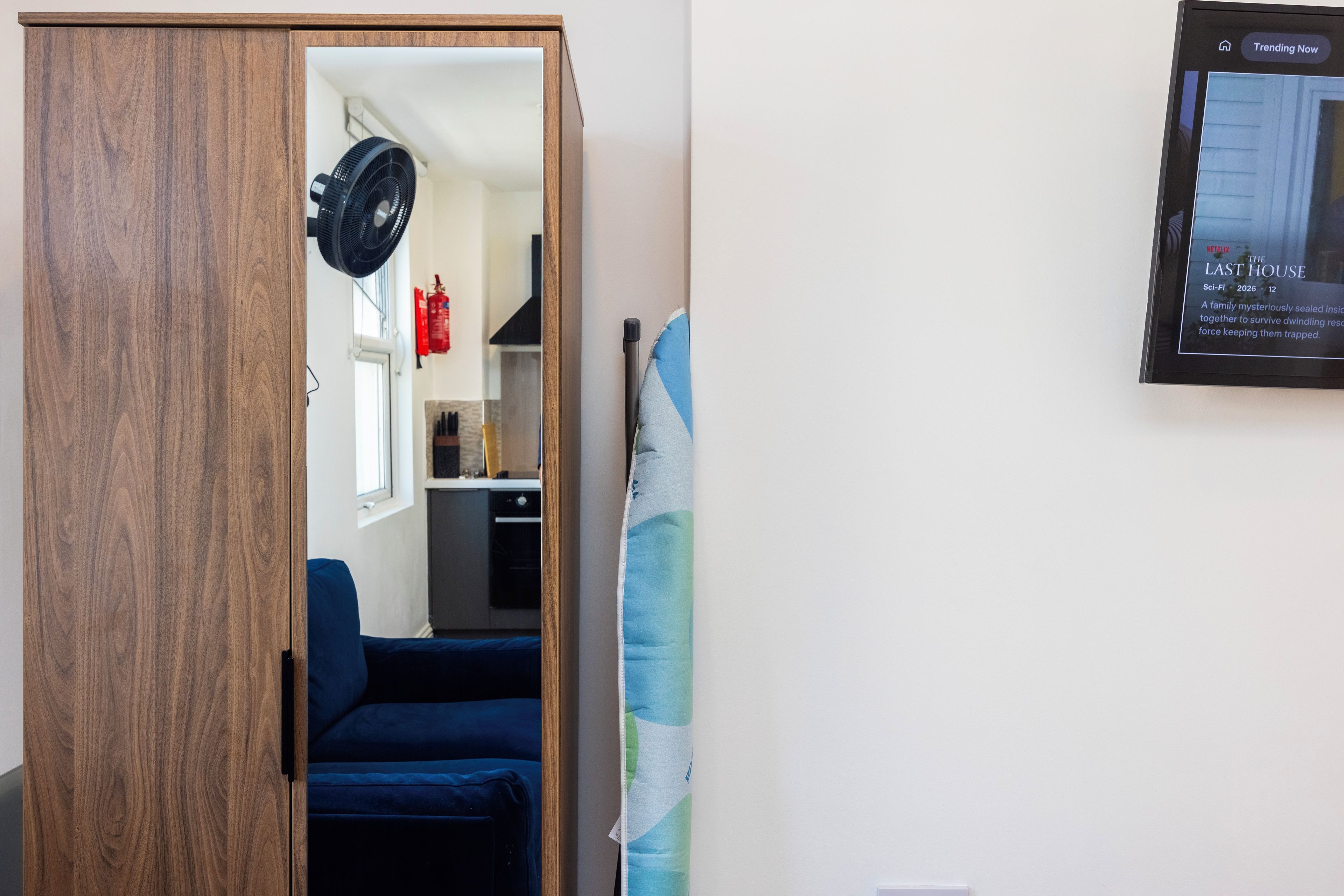 A room with a wooden wardrobe, a blue sofa, a standing fan, and a framed picture on the wall.