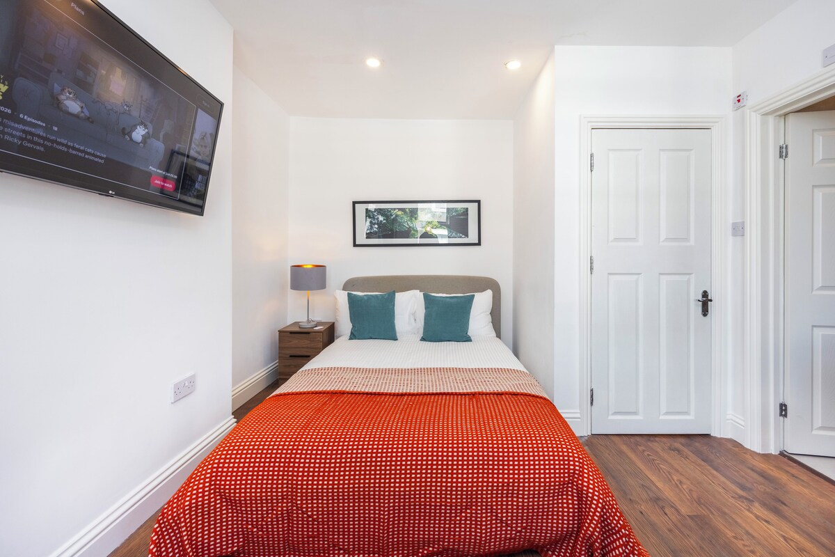 A bedroom with a bed, a TV mounted on the wall, a framed picture above the bed, and a door to the right.