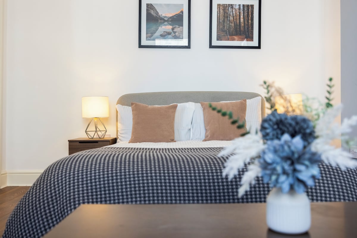 A neatly made bed with a checkered bedspread and pillows, a bedside table with a lamp, and two framed pictures on the wall.