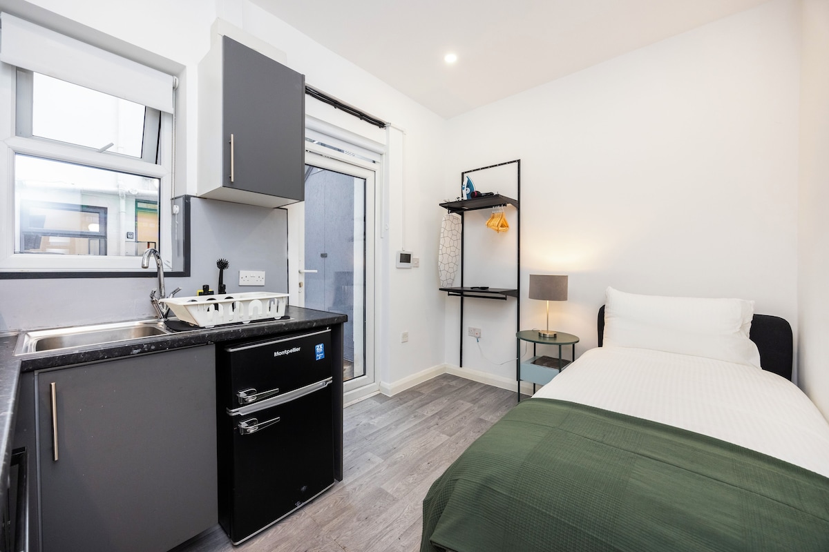 A compact hotel room with a kitchenette, a bed, a bedside table, and a small desk.