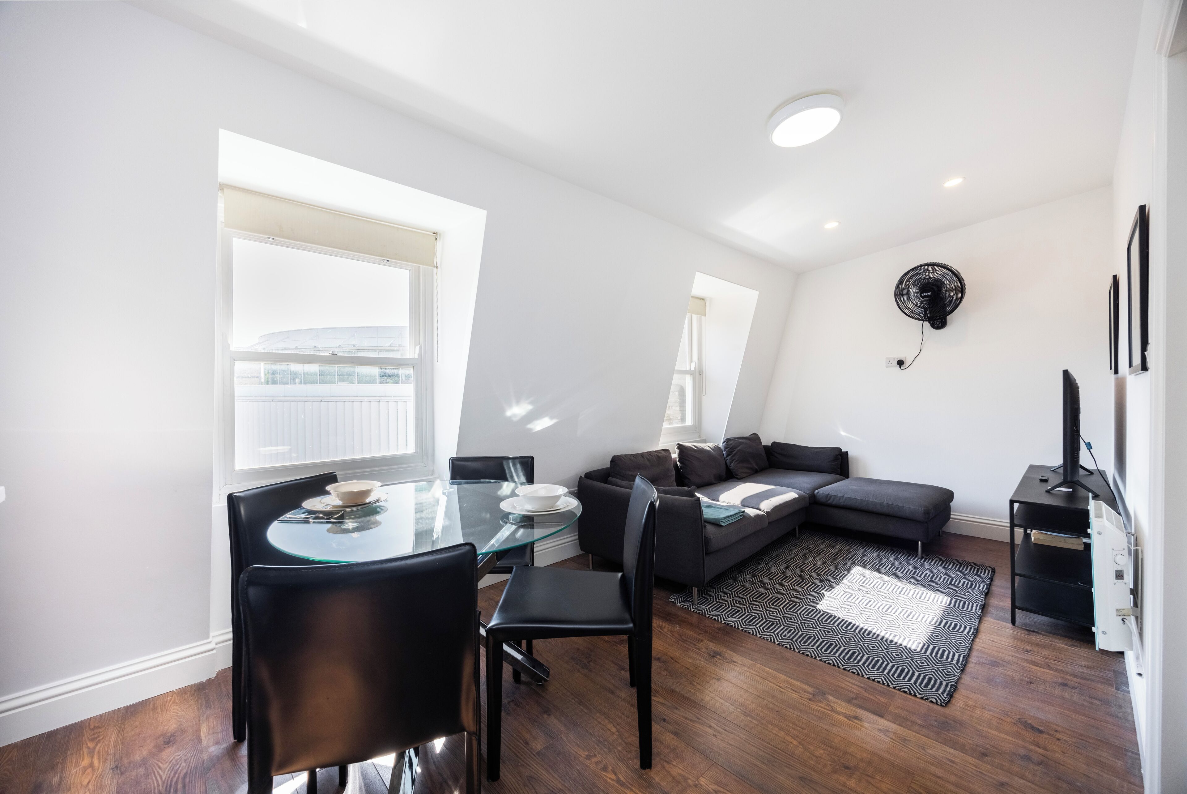 A modern living space with a glass dining table, black leather chairs, a gray sofa, and a patterned rug.