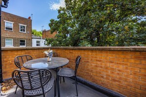A patio with a round table and four chairs, a brick wall, and a tree in the background. - Earl's Court Residence (London)