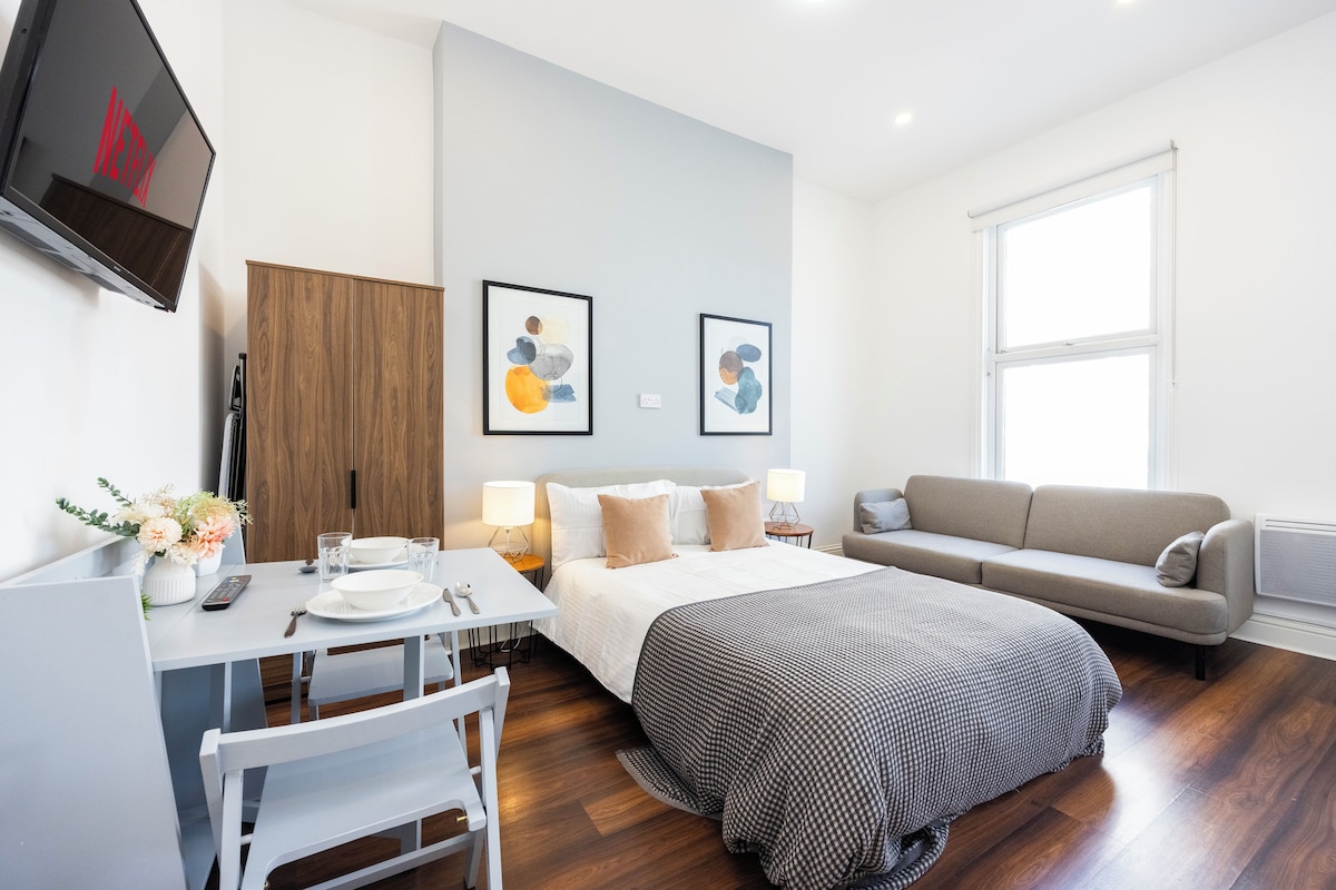 A modern hotel room with a bed, a sofa, a desk with a chair, a TV mounted on the wall, and two framed artworks.