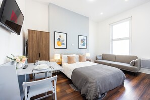 Habitación de hotel moderna con una cama, un sofá, un escritorio con una silla, un televisor montado en la pared y dos cuadros enmarcados.