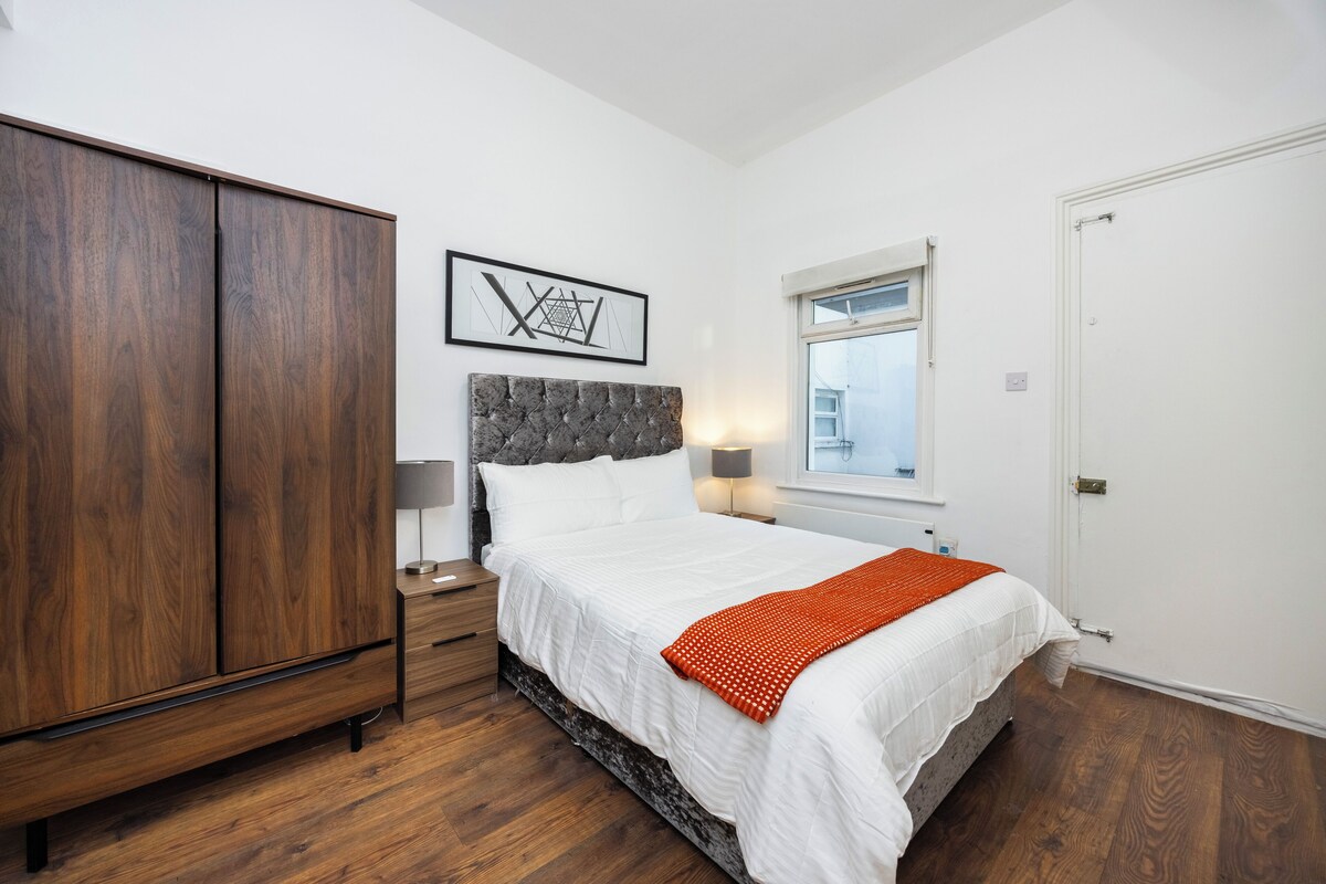 A bedroom with a bed, a wardrobe, a nightstand, a lamp, a wall art piece, and a window.