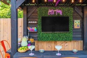 An outdoor bar area with a neon sign, a TV screen, and a wooden fence. - Heated Pool, HotTub, Pickleball, Golf & Game Room (San Antonio)