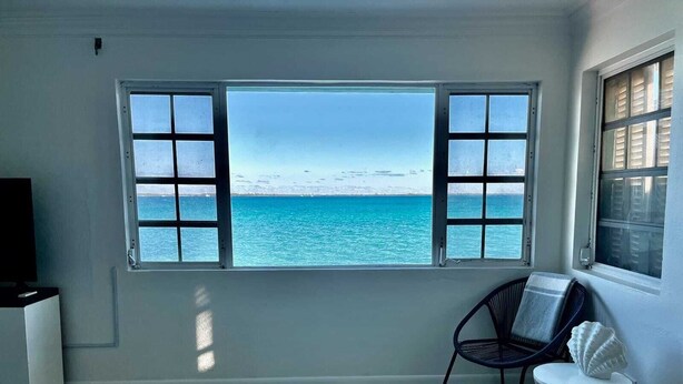 Ein Zimmer mit großem Fenster und Meerblick, ein Sessel, ein kleiner runder Tisch und ein Fernseher auf einem Schrank.