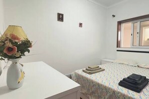 A bedroom with a bed, a bedside table with a lamp and a vase of flowers, a window with blinds, and a small picture on the wall. - Charming 3-bedroom House Rental in Trofa, Portugal (Trofa)