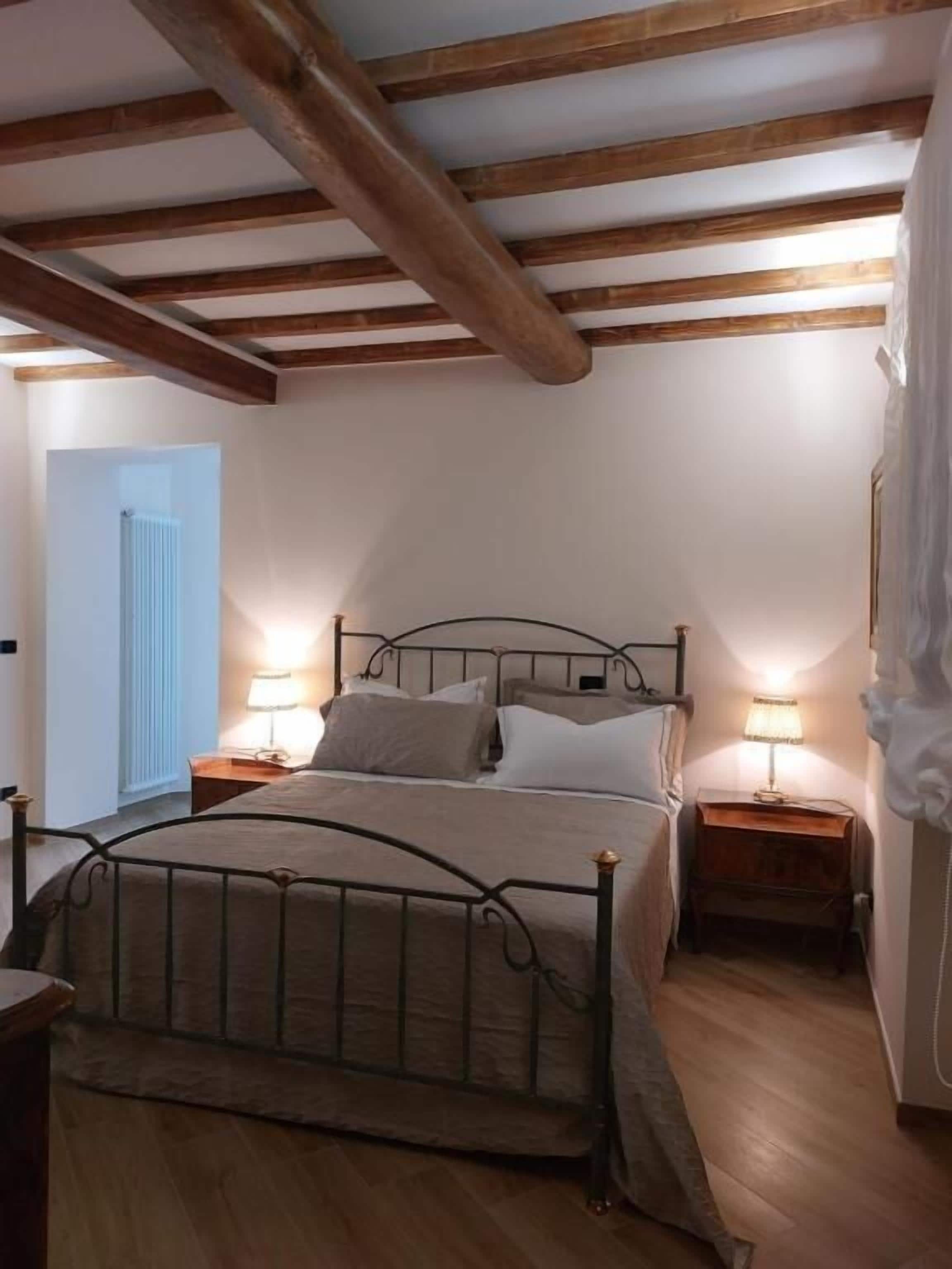 Un dormitorio con estructura de cama metálica, dos mesitas de noche con lámparas, suelo de madera y techo inclinado con vigas a la vista.