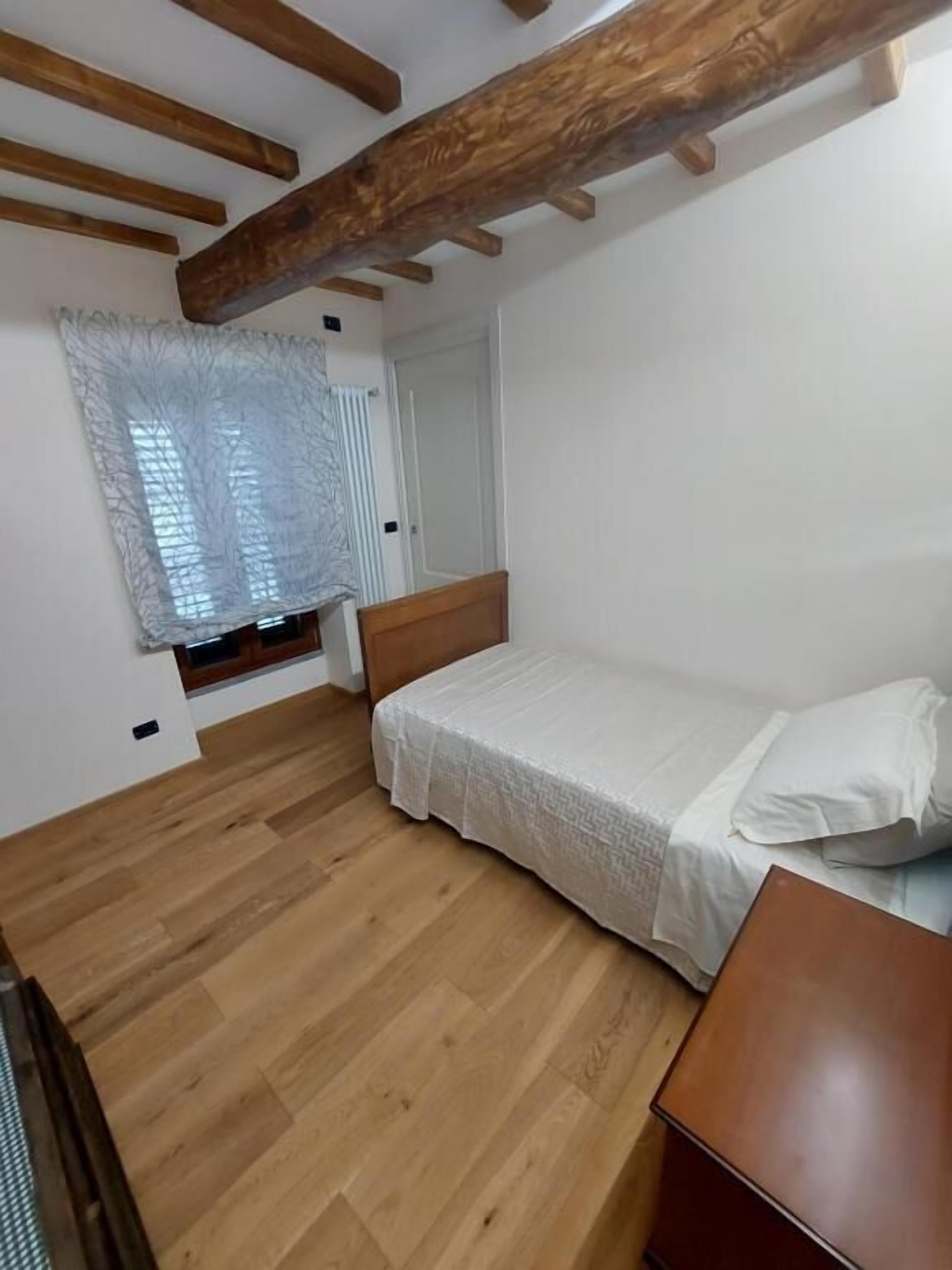 A bedroom with a bed, wooden flooring, a window with a white curtain, and a door.
