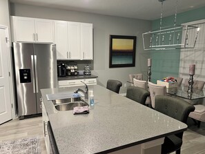 A modern kitchen with a stainless steel refrigerator, a coffee station, and a dining area with a glass table and cushioned chairs. - Spacious 4-bedroom house with pool and gym access not far from beaches! (Palmetto)
