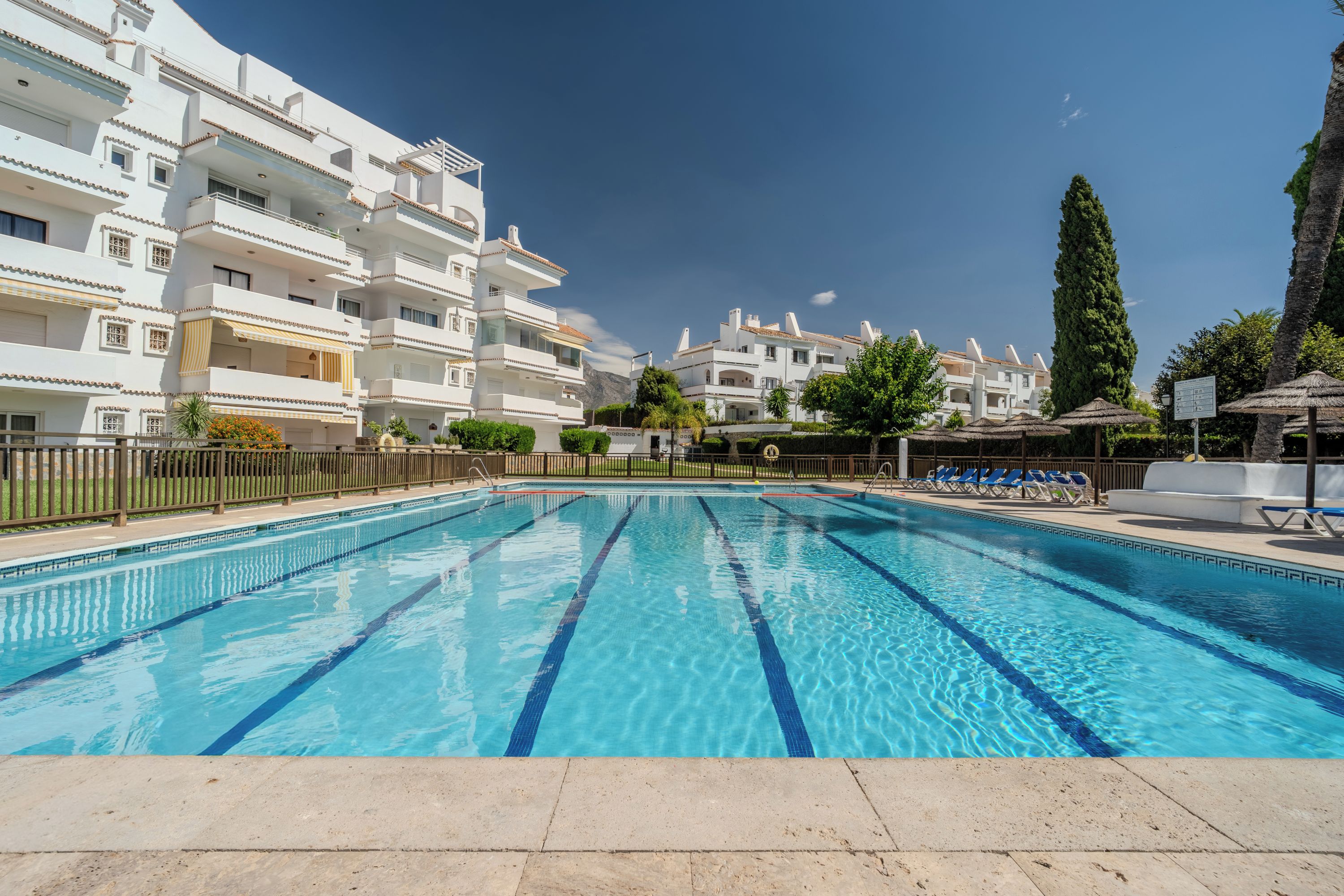 A swimming pool with clear blue water, surrounded by a fence, with a view of white buildings in the background.