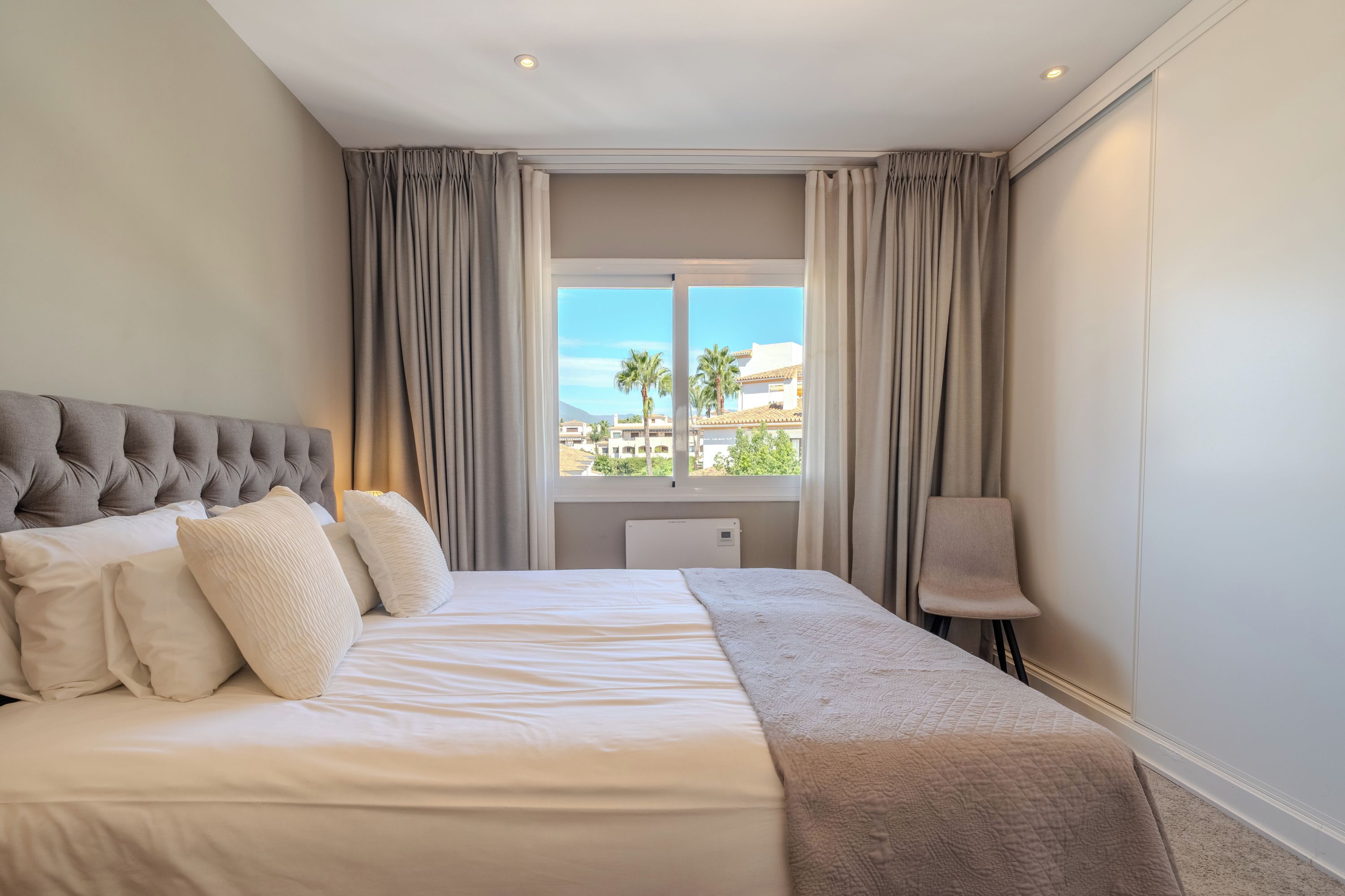 Habitación de hotel con cama, una silla, ventana con cortinas y vista a edificios y palmeras.