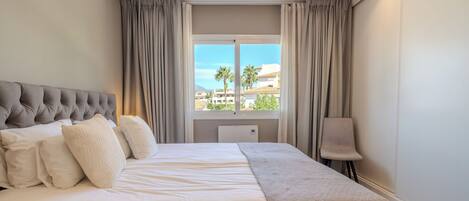 Habitación de hotel con cama, una silla, ventana con cortinas y vista a edificios y palmeras.