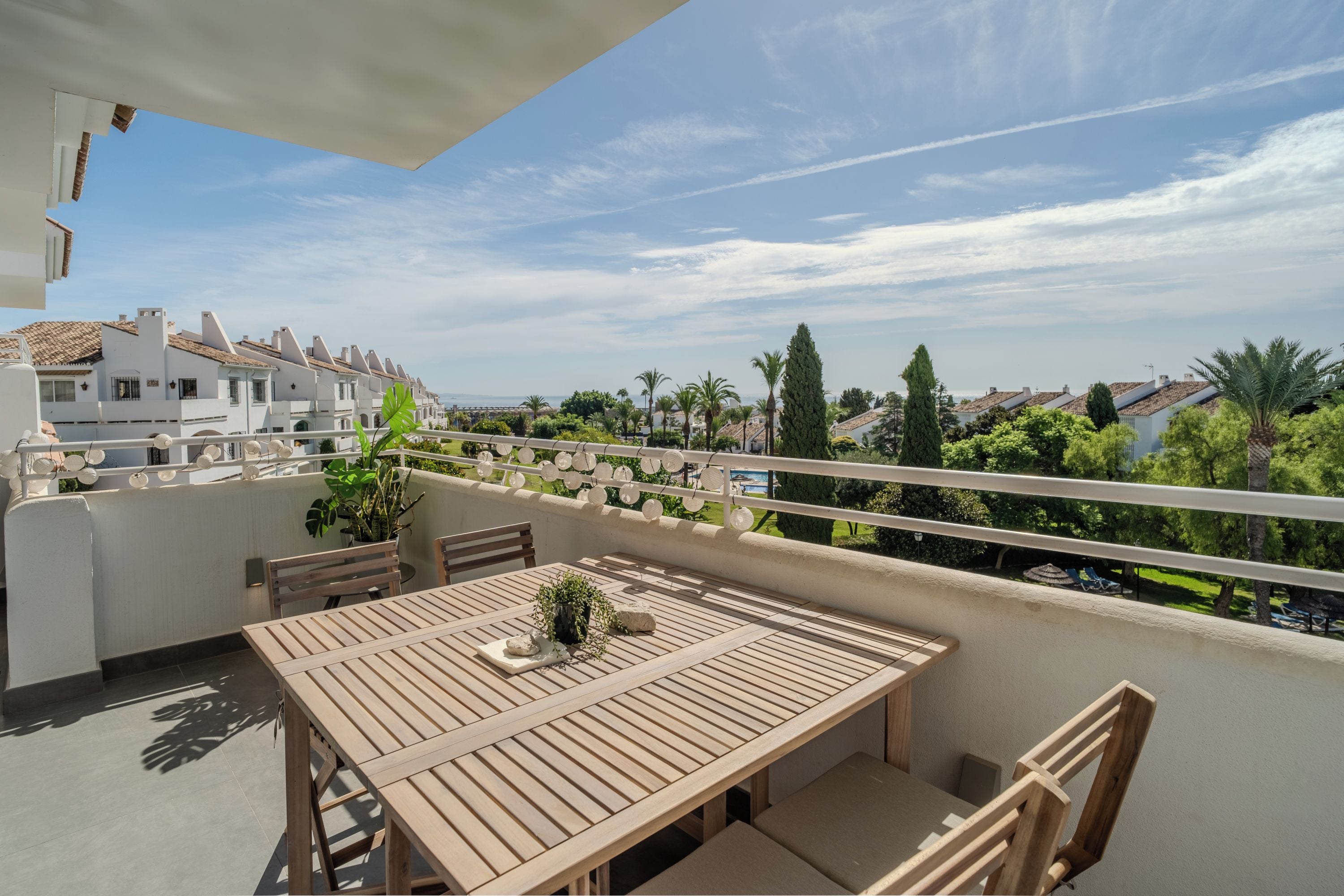 Una mesa y sillas de madera en un balcón con vista a una zona residencial y un cielo despejado.