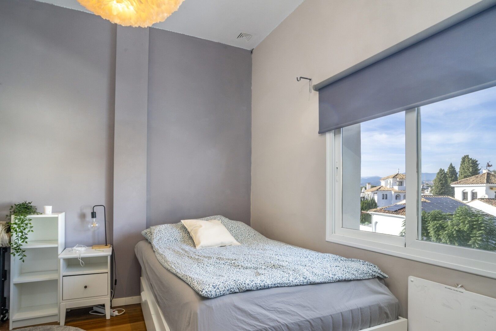 Een slaapkamer met een bed, nachtkastje, lamp, raam met uitzicht op gebouwen, en een radiator.