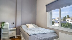 A bedroom with a bed, a bedside table, a lamp, a window with a view of buildings, and a radiator. - Rg133 - Spacious 3 bed Apartment in Prime Location (Marbella)