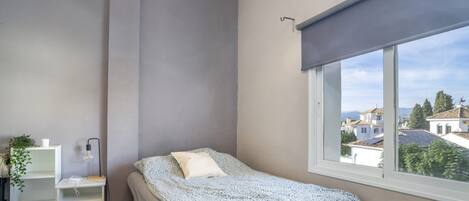 Een slaapkamer met een bed, nachtkastje, lamp, raam met uitzicht op gebouwen, en een radiator.