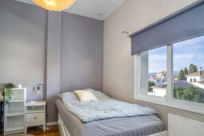 Ein Schlafzimmer mit Bett, Nachttisch, Lampe, Fenster mit Blick auf Gebäude und Heizkörper.