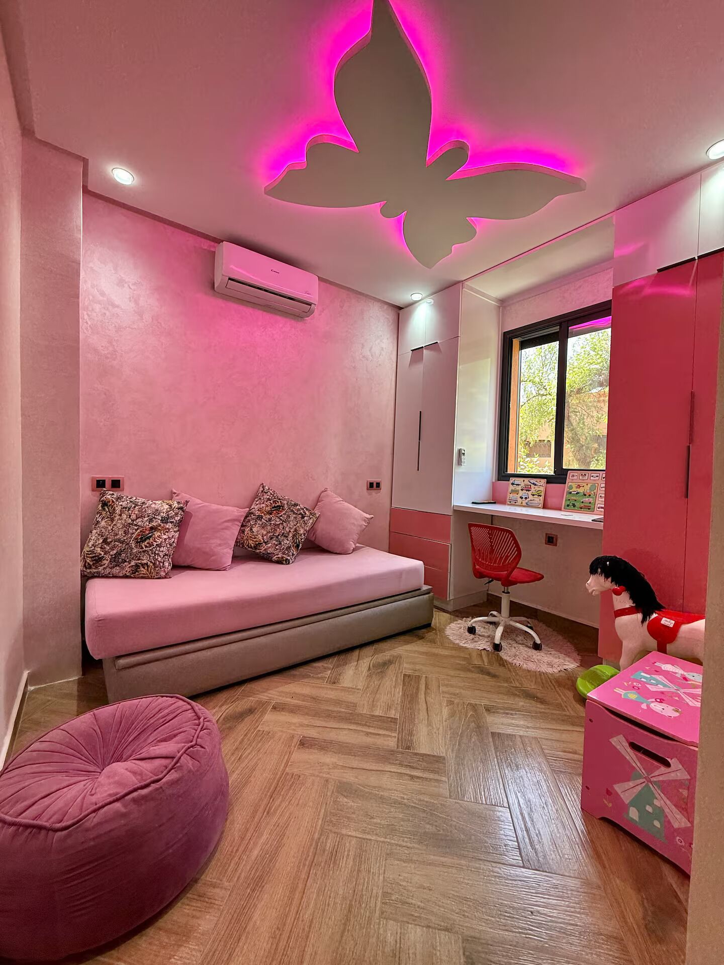 A pink-themed room with a sofa, a bean bag, a desk with a chair, and a window.