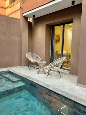 Un espace aménagé au bord de la piscine, avec deux fauteuils en osier blanc et une petite table, une véranda fermée par des baies vitrées, et une vue sur l’intérieur à travers une porte en verre.
