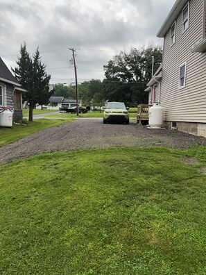 A residential driveway with a green car parked on the side, a large white propane tank, and a house with a visible chimney.