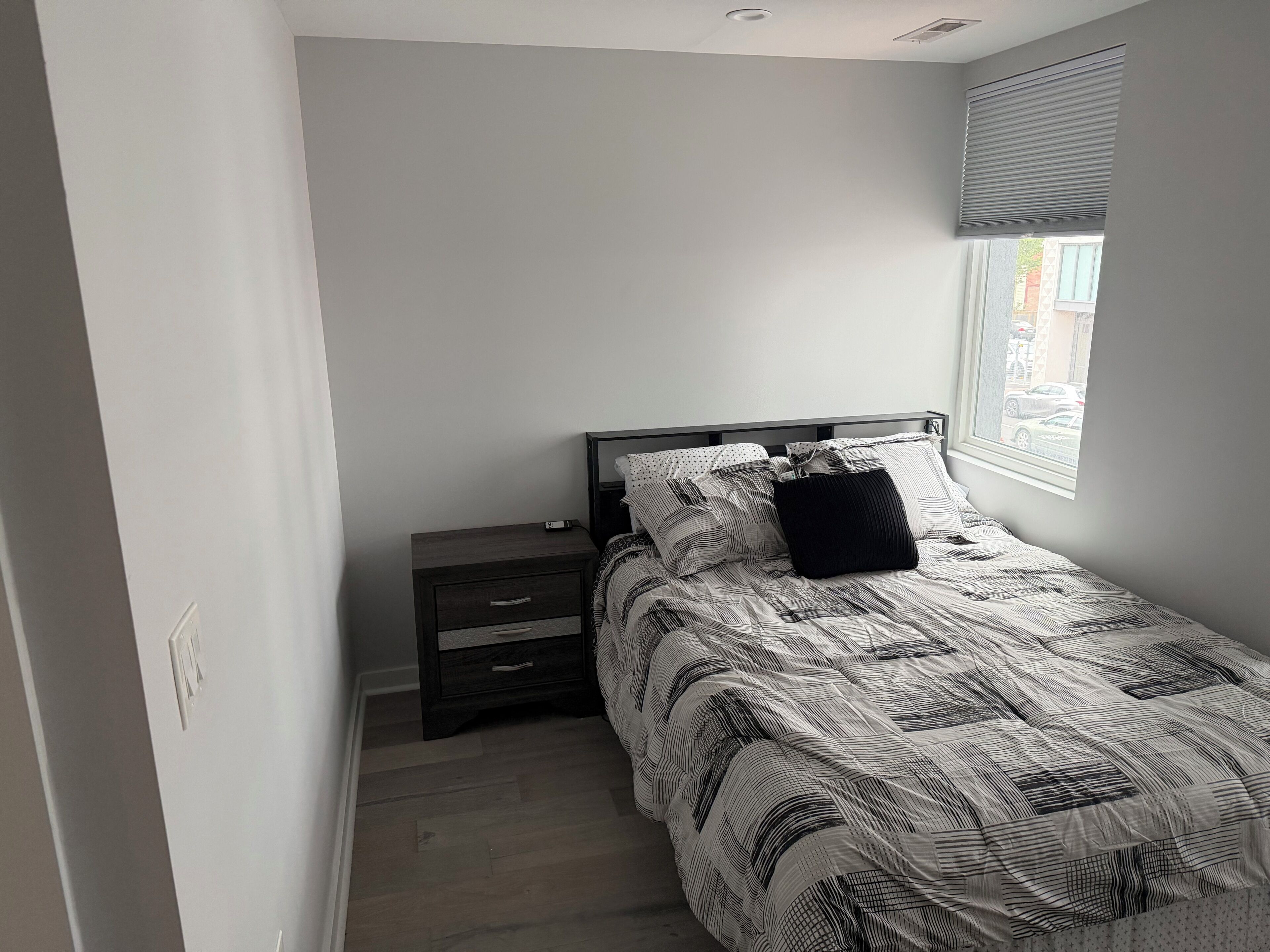 A bedroom with a bed, a nightstand, a drawer, a window with blinds, and a view of the street.