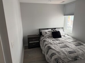 Ein Schlafzimmer mit einem Bett, einem Nachttisch, einer Schublade, einem Fenster mit Jalousien und Blick auf die Straße.