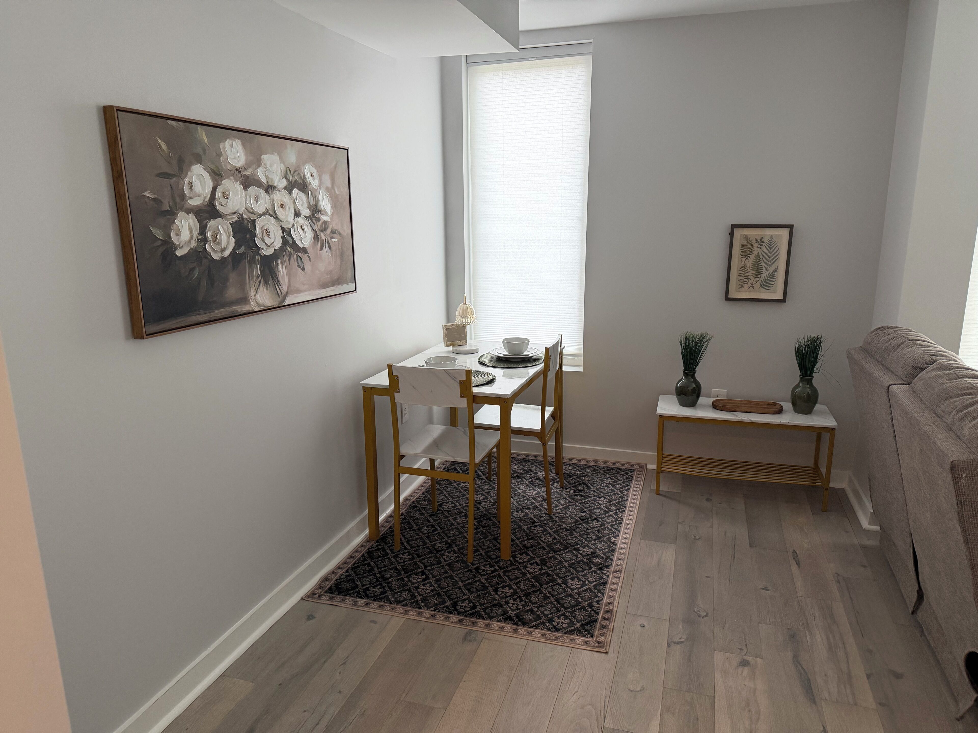 A modern living room with a dining area, featuring a white table, wooden chairs, a patterned rug, and a grey sofa.