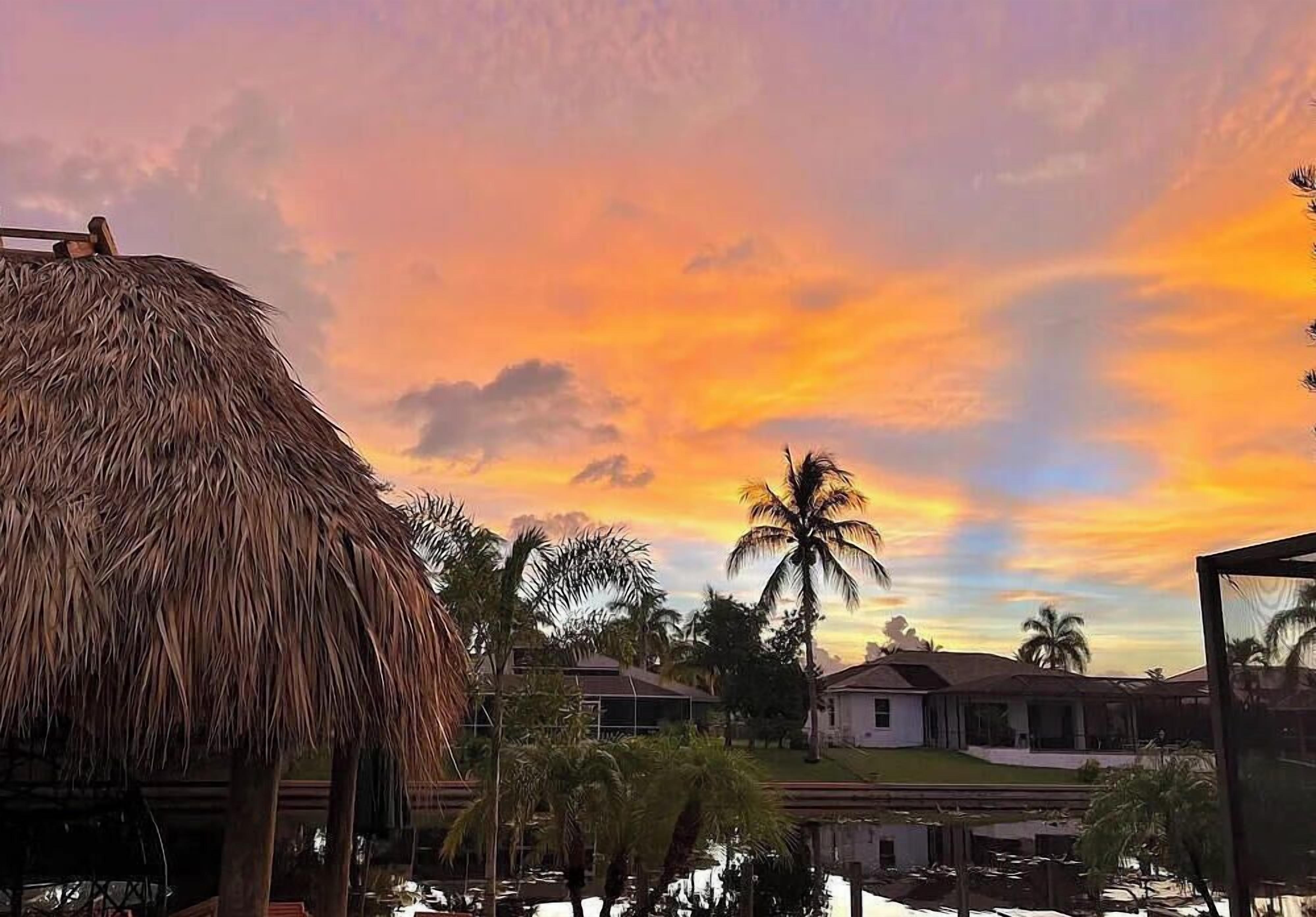 A thatched-roof structure, palm trees, and houses against a sunset sky.
