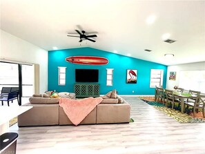 Ein modernes Wohnzimmer mit einer türkisfarbenen Akzentwand, Deckenventilator, Flachbildfernseher und einem Essbereich mit Holzsesseln und einem Tisch.