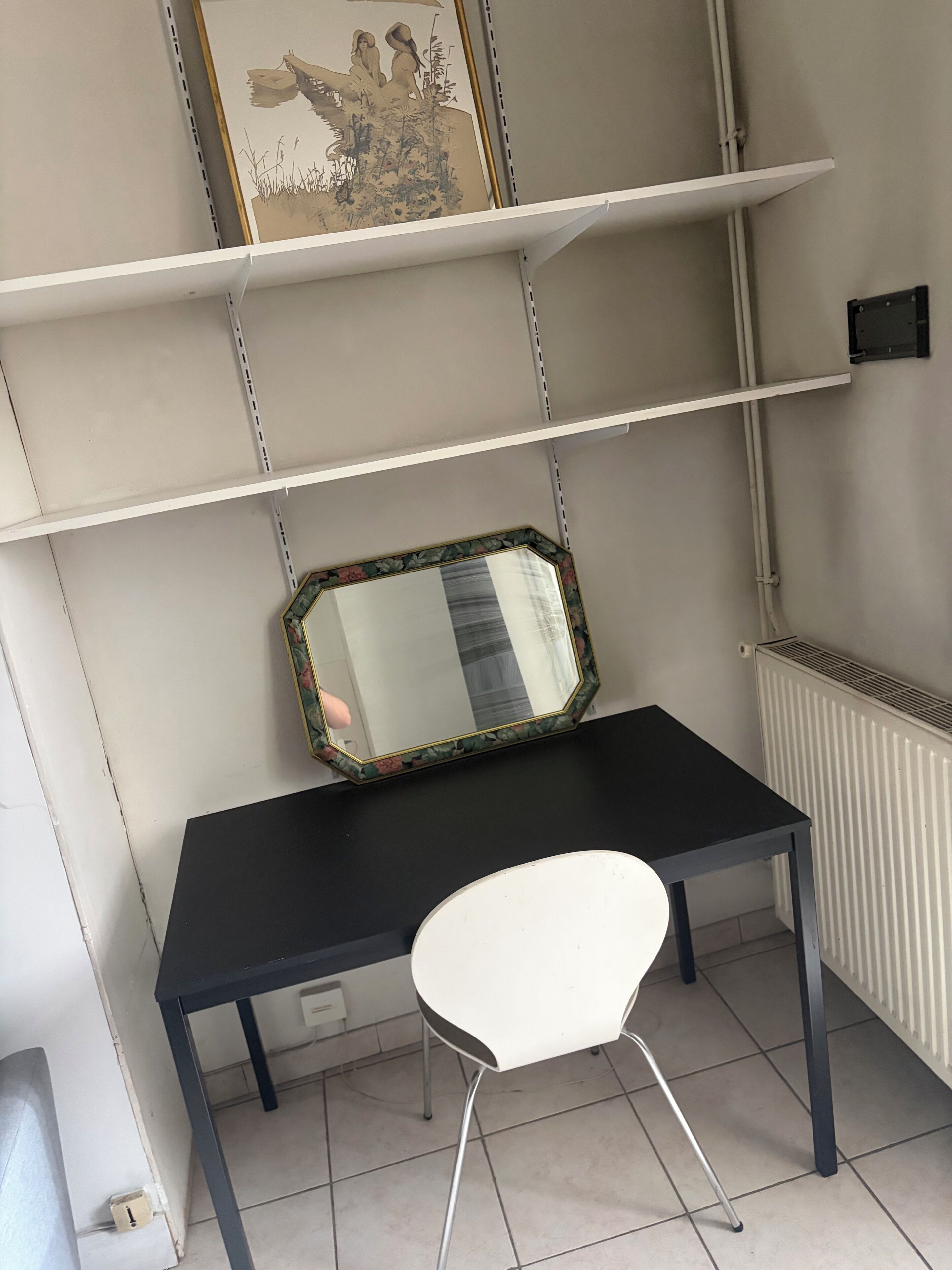 Una habitación con un escritorio negro, una silla blanca, un cuadro en la pared y un radiador.