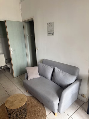 Ein Zimmer mit einem grauen Sofa, einem kleinen runden Holztisch und einer Tür zu einem Badezimmer.
