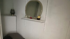 Quarto pequeno com uma cama de solteiro, colchão e travesseiro brancos, mesa de cabeceira com abajur e um espelho na parede.