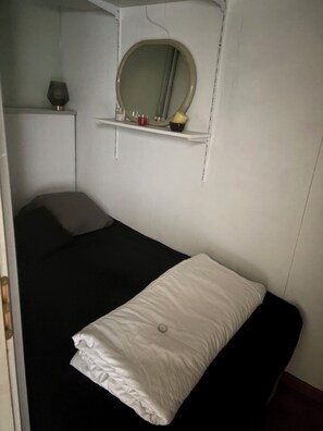 Ein kleines Zimmer mit einem Einzelbett, einer weißen Matratze und einem Kopfkissen, einem Nachttisch mit einer Lampe und einem Spiegel an der Wand.