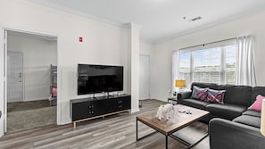 A modern living room with a flat-screen TV, a sofa, a coffee table, and a window with blinds. - Roll Tide in Luxury - Premium 2BR Ultimate Bama Getaway (Tuscaloosa)