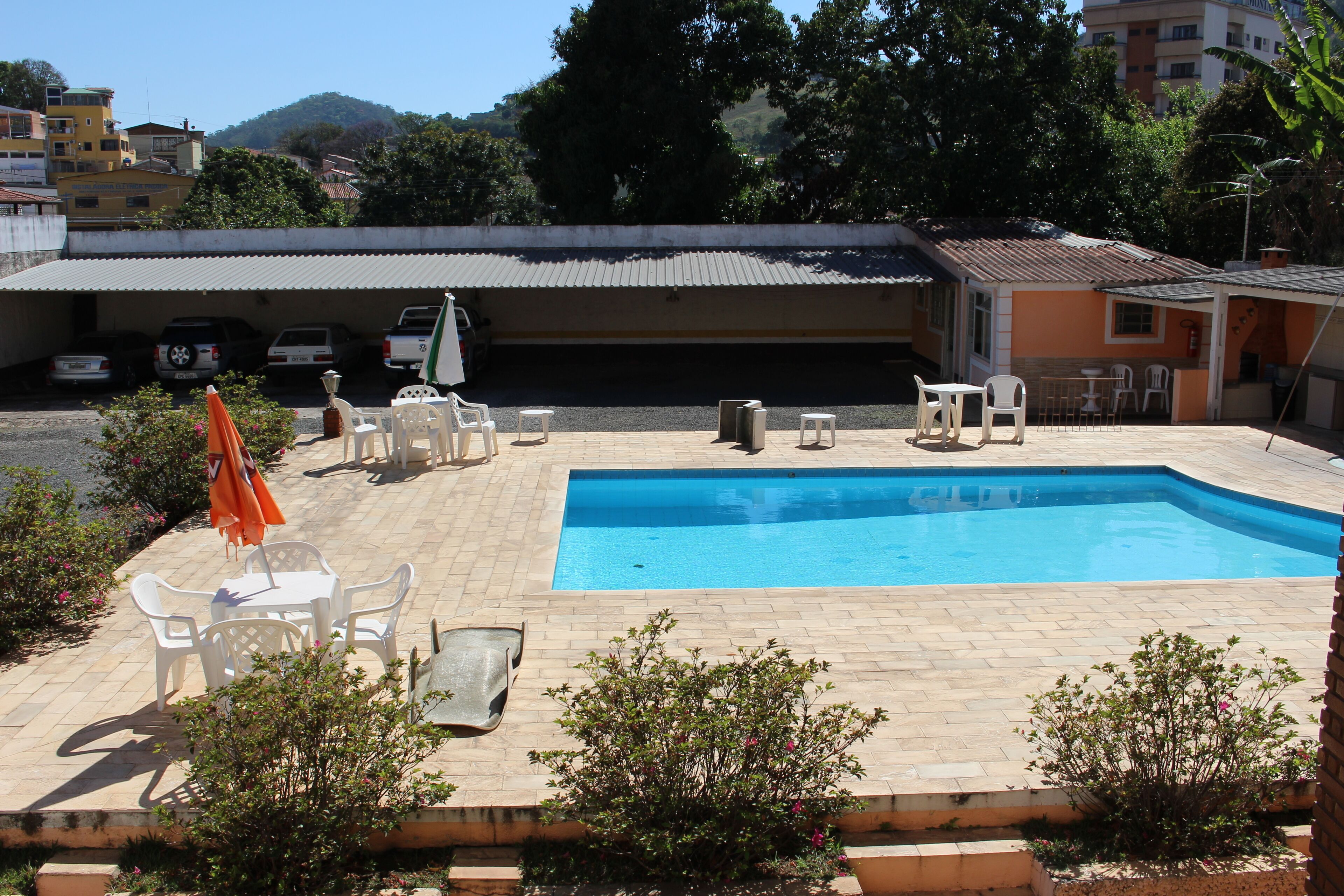 Una piscina con borde de baldosas, rodeada de sillas y mesas de plástico blanco, y un edificio al fondo.