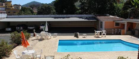 Una piscina con borde de baldosas, rodeada de sillas y mesas de plástico blanco, y un edificio al fondo.