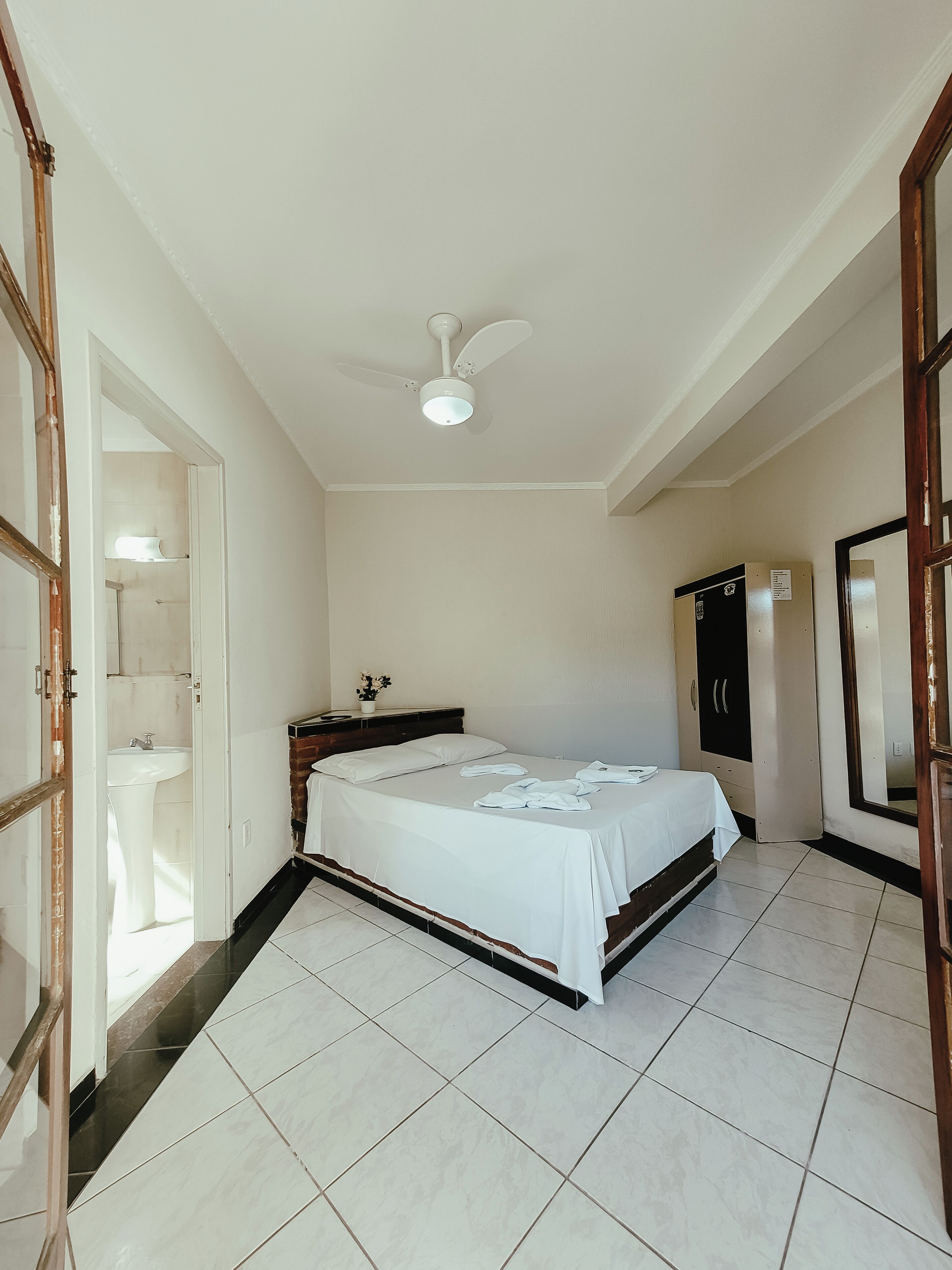 Habitación de hotel con cama, ventilador de techo, armario y baño visible a través de una puerta abierta.