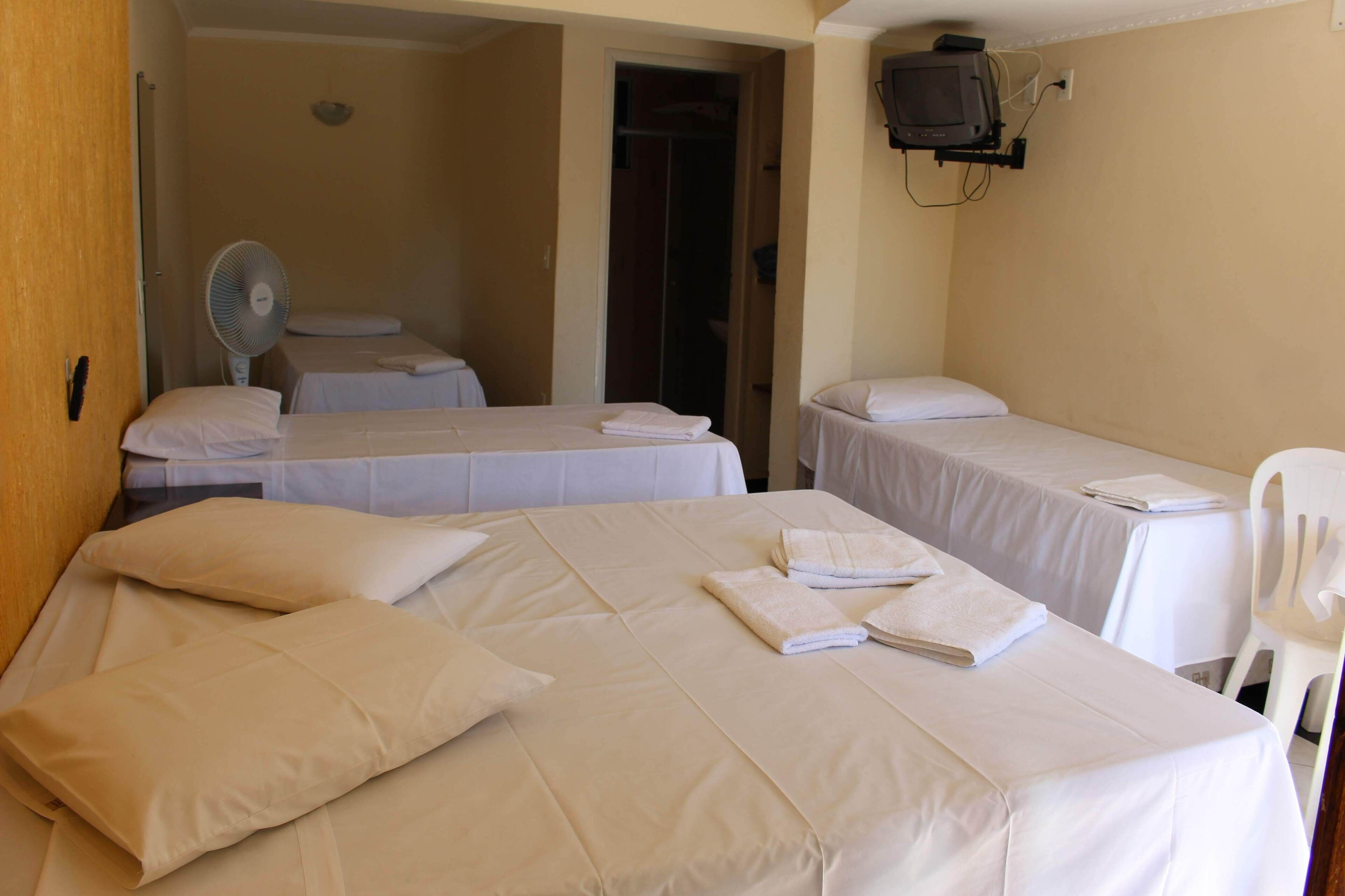 A hotel room with two beds, each with white linens and pillows, a single bed with a white chair, and a TV mounted on the wall.