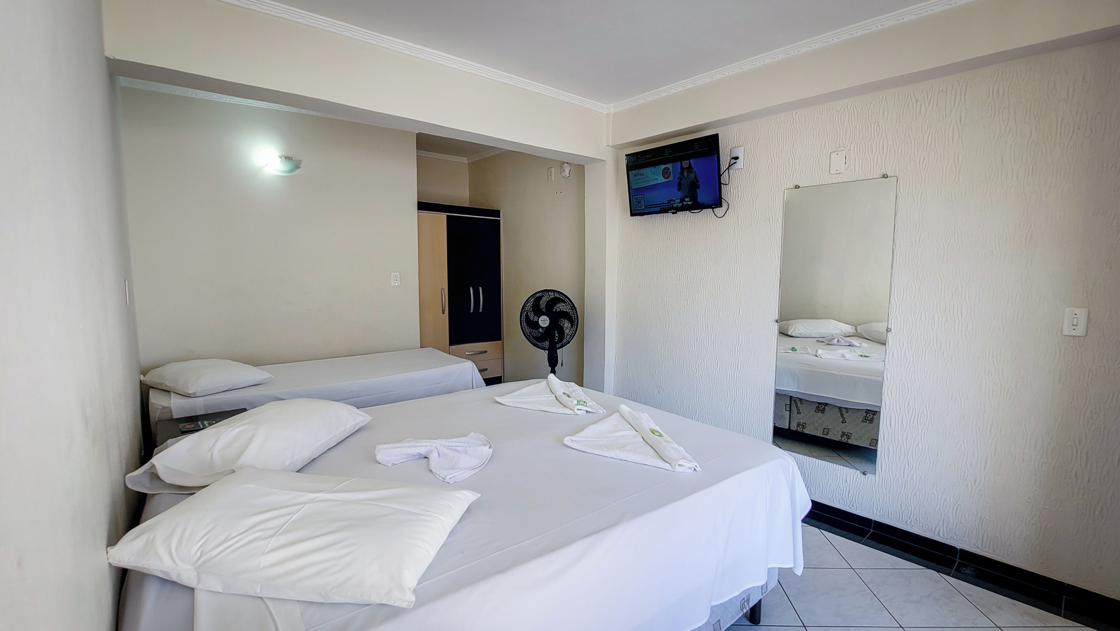 Quarto de hotel com duas camas, televisão fixada na parede, um ventilador e um espelho.