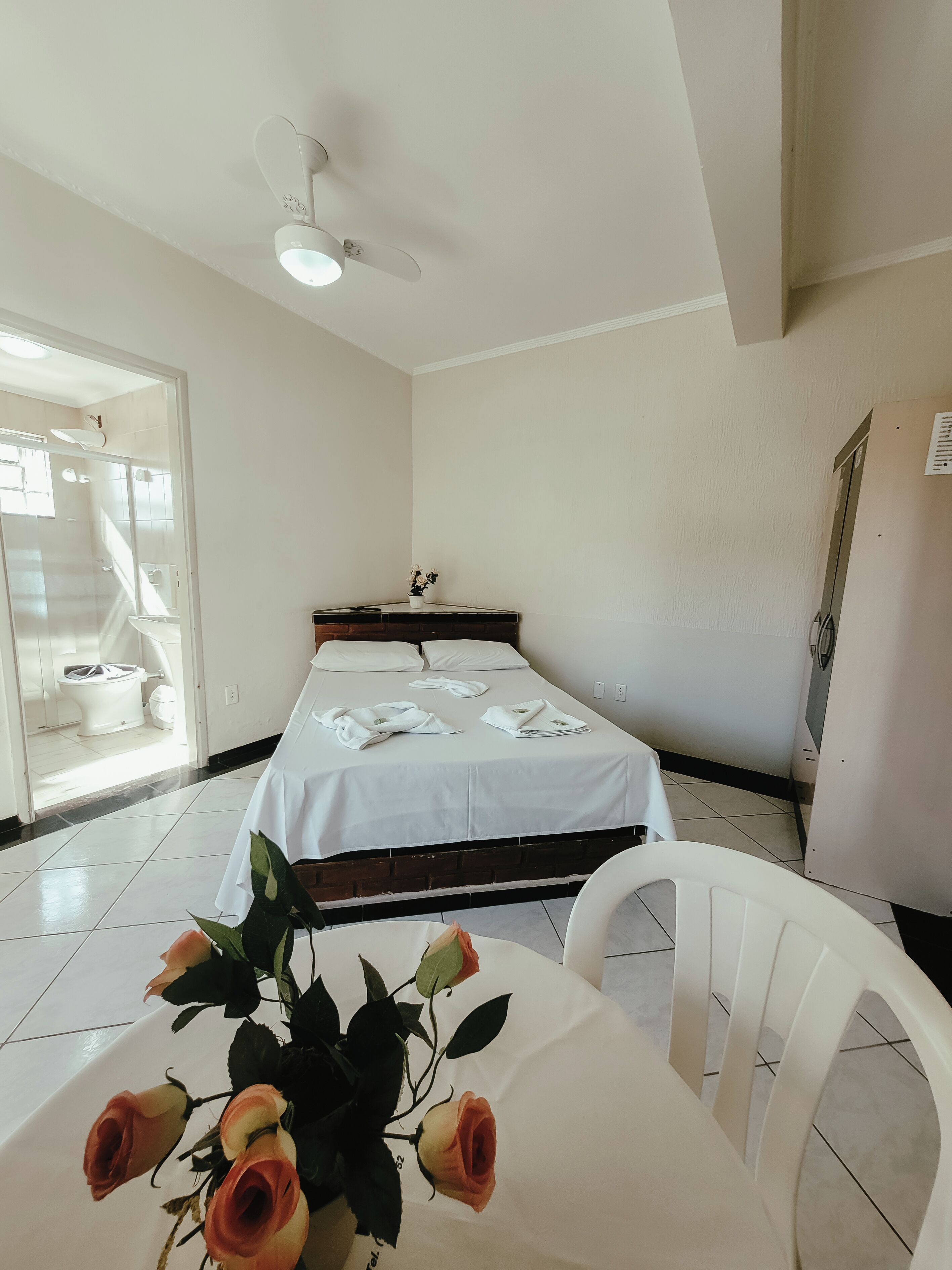 Habitación de hotel con cama, ventilador de techo, baño visible a través de una puerta abierta y una mesa con un jarrón de rosas.